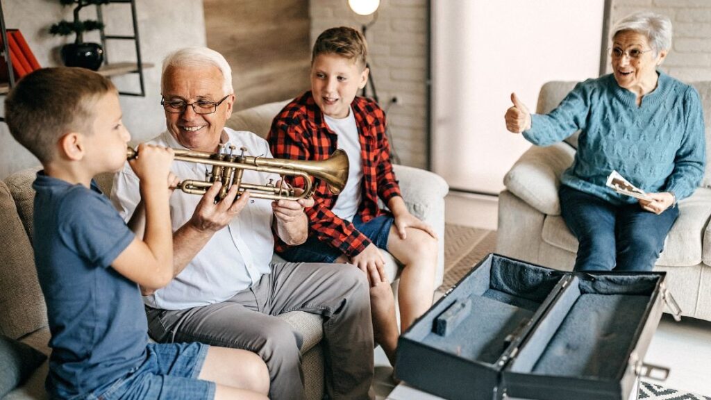 young boy blowing into a trumpet with another boy and older adults looking on and helping