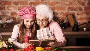 Older woman with young girl in a cook's hat looking at an old recipe book.