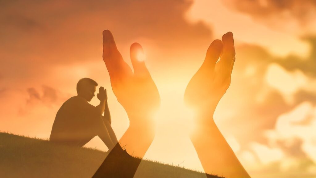 A man sitting with hands held in prayer on a hilside against a brilliant sunlight sky. A set of hands superimposed reaching for the sun.