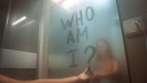 Woman writing "who am I?" in the bathroom mirror.