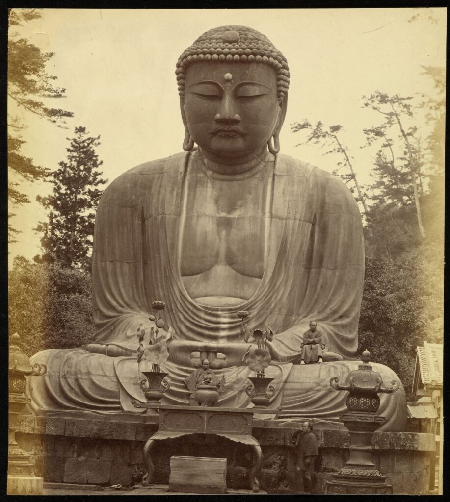 vintage photograph of bronze statue of a Great Buddha (Daibutsu) at the temple of Kotoku-in. Two men sit at the base of the statute, another stands by the stone pedestal.