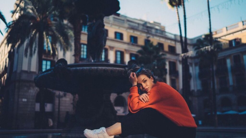 woman sitting in front of a building with a fountain and palm trees looking discouraged 