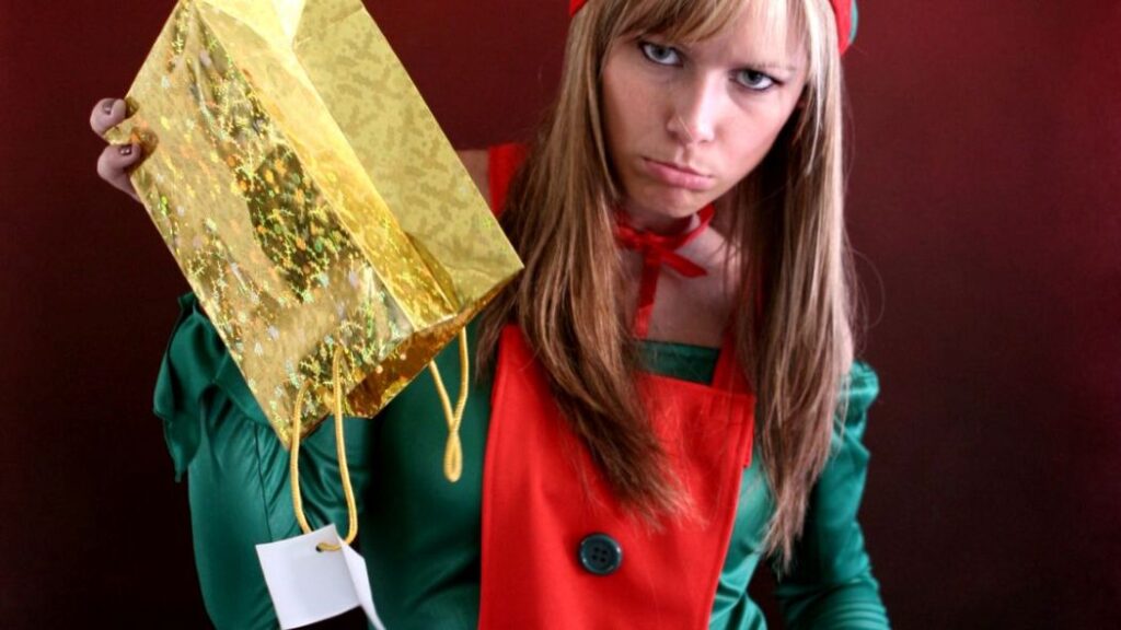 woman dressed as a christmas elf holding a holiday bag upside down and looking disappointed because it is empty