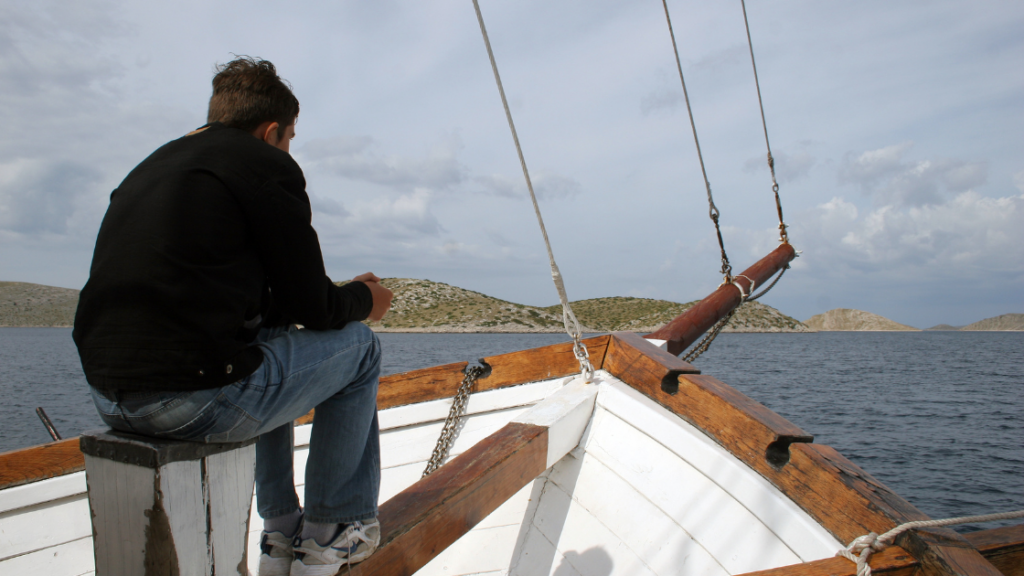 man sitting on the bow of a sailboat staring out at the water and land in the distance